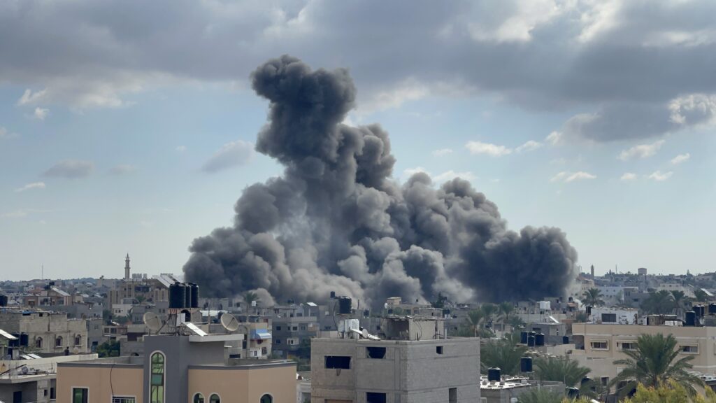 A large column of dark grey smoke rises between densely packed civilian apartment buildings in Gaza during morning airstrikes. The smoke plume dominates the centre of the frame, dwarfing the surrounding residential towers. The image captures both the immediate violence of the attack and the atmospheric emissions it produces.